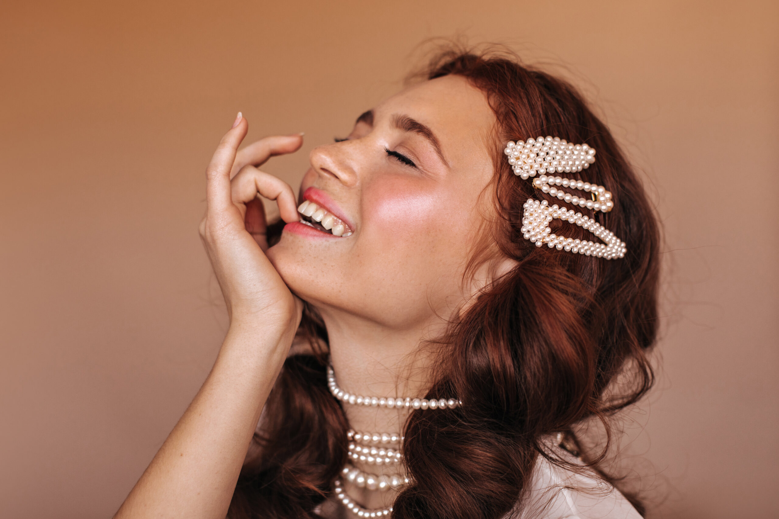 Positive girl with curly hair laughs and bites her finger. Portrait of woman with white hairpins and pearl necklace